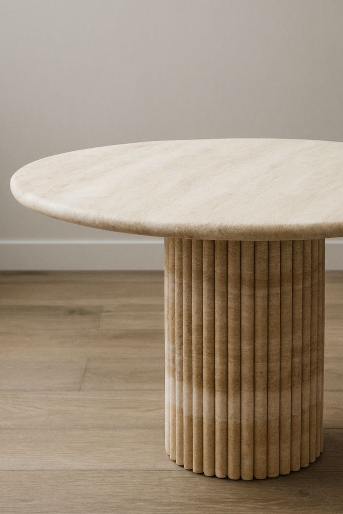 Round travertine table with a ribbed cylindrical base on a wooden floor.